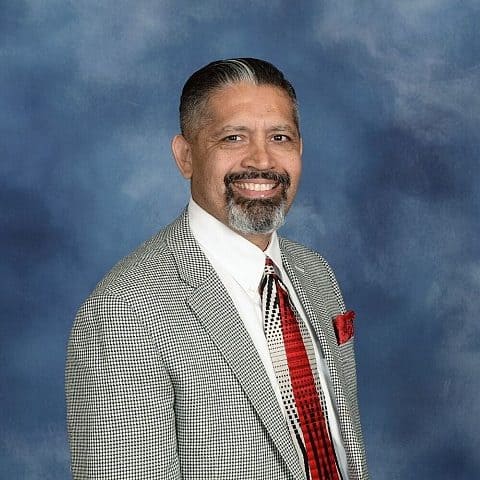 Gene Roque, with a salt-and-pepper beard wearing a houndstooth blazer and red tie, smiling against a blue mottled background.