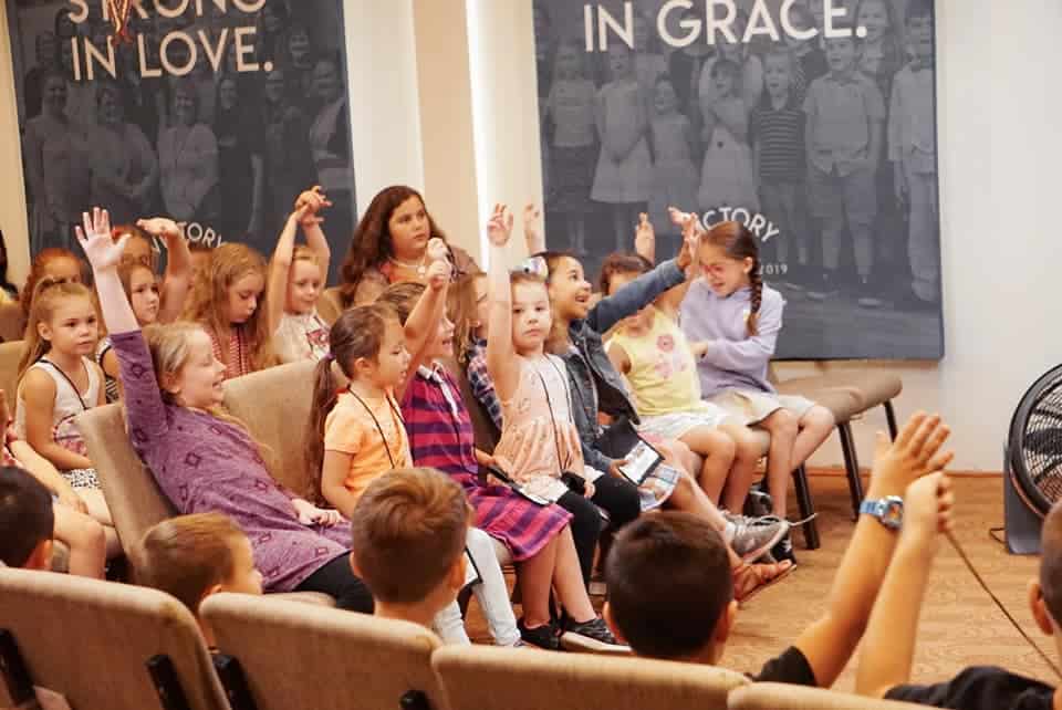 A group of children raising their hands during a session in a room with adults supervising, part of the Kid's Ministry in a church.