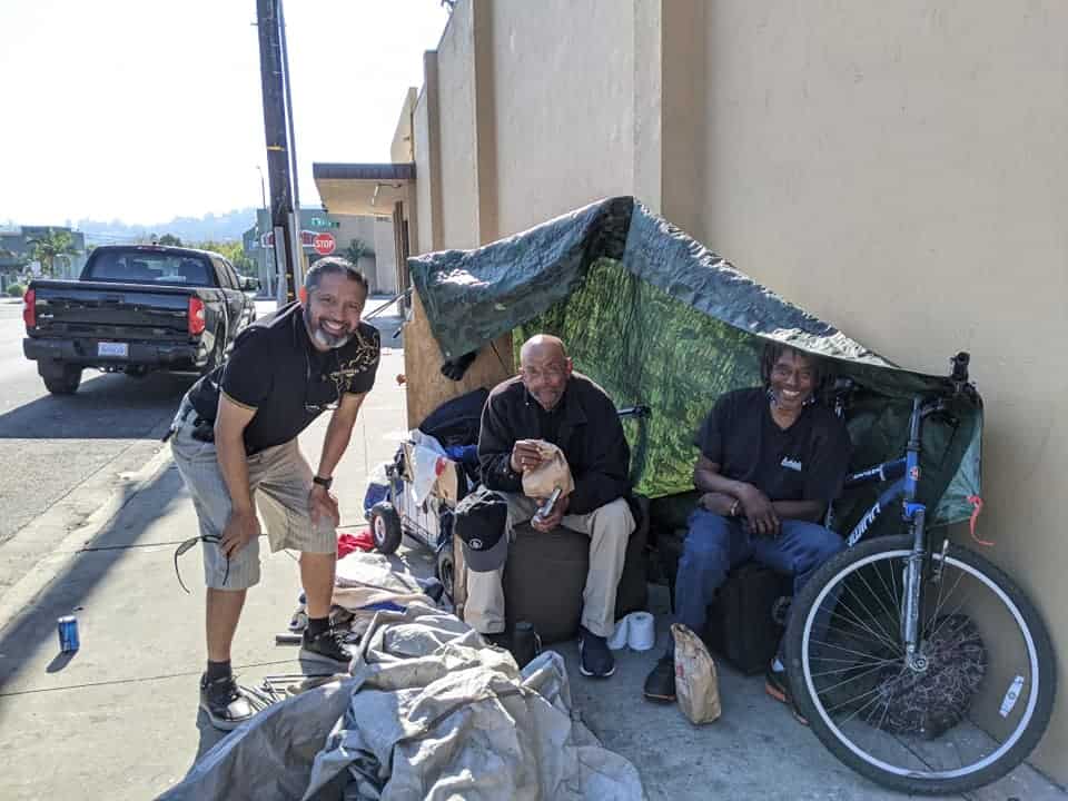 Three individuals, two sitting and one standing, beside a makeshift shelter on a street, with a bicycle and belongings, smiling towards the camera during a Homeless Outreach event.