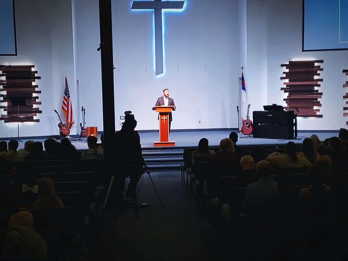 Man speaking at a podium in a church auditorium, audience in the foreground, with a large cross illuminated on the wall behind, as part of a Church Service.