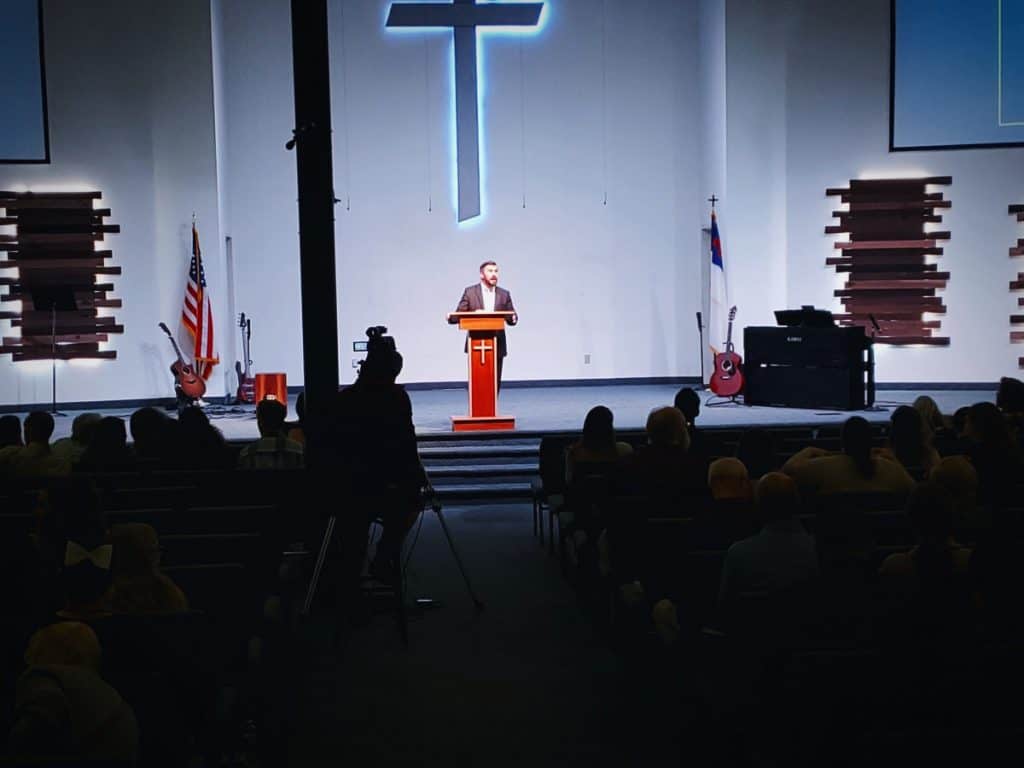A man speaks at a podium in a church auditorium, with a large cross on the wall behind him and an audience listening, at Victory Baptist Church in El Cajon.