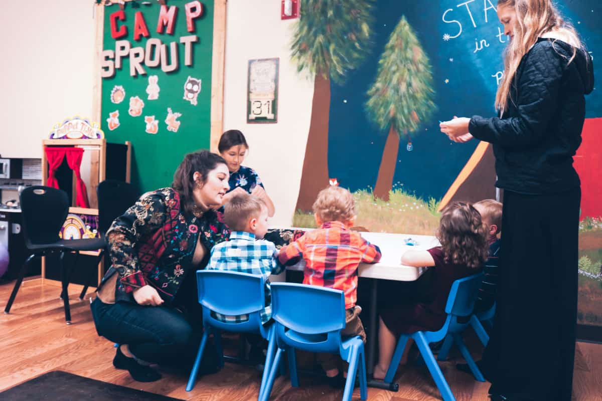In a vibrant classroom setting filled with decorations, a teacher and volunteer engage with young children seated at a table, while another adult oversees the activity in the background, all part of the dynamic Kid's Ministry.