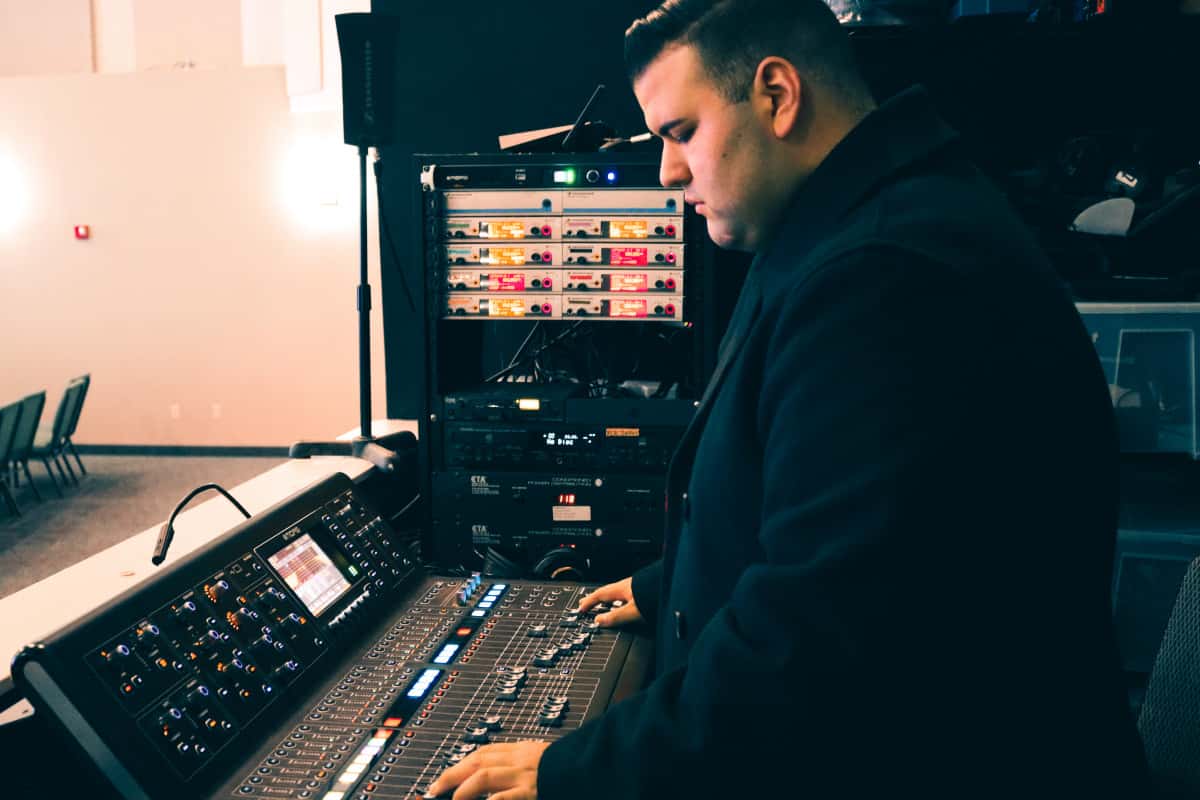 A volunteer adjusts settings on a large audio mixing console in a dimly lit room, part of the "Sound Ministry."