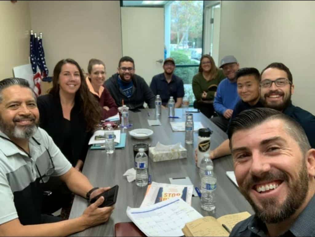A group of people smiling at a conference table during a meeting, with snacks and water bottles visible, part of a "Staff Meeting."