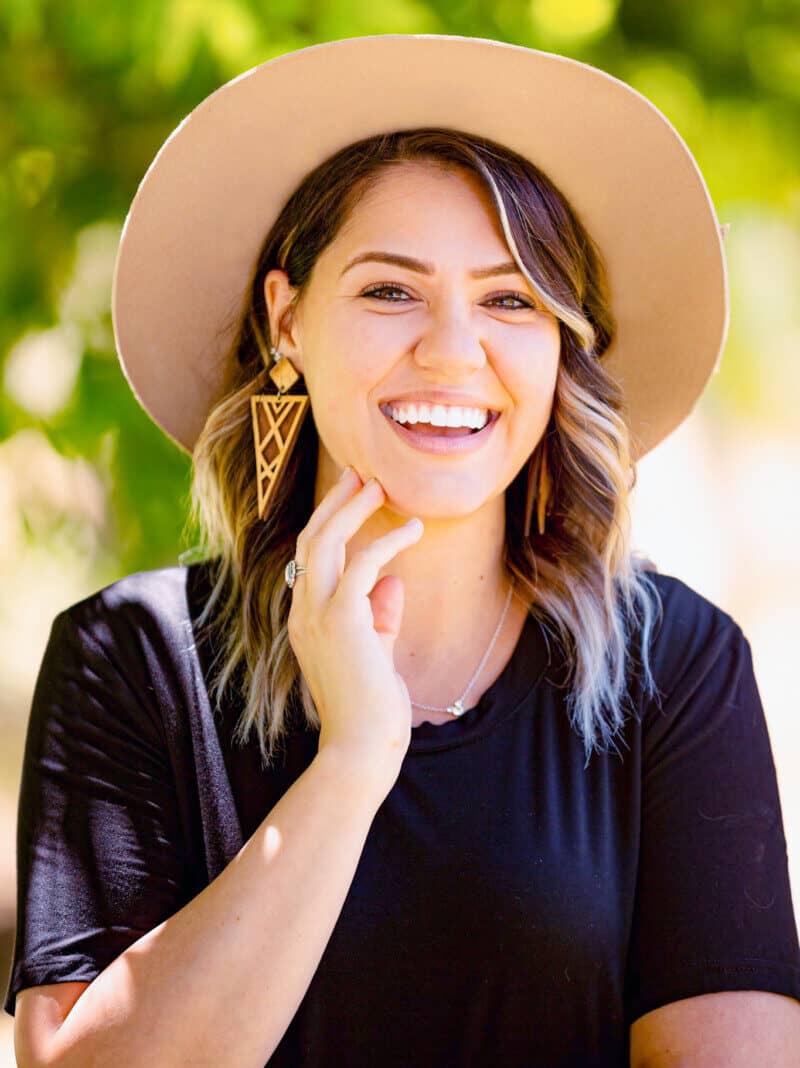 A woman with wavy hair, wearing a wide-brimmed hat and geometric earrings, smiles brightly. She touches her chin with one hand and wears a black shirt amidst blurred greenery.