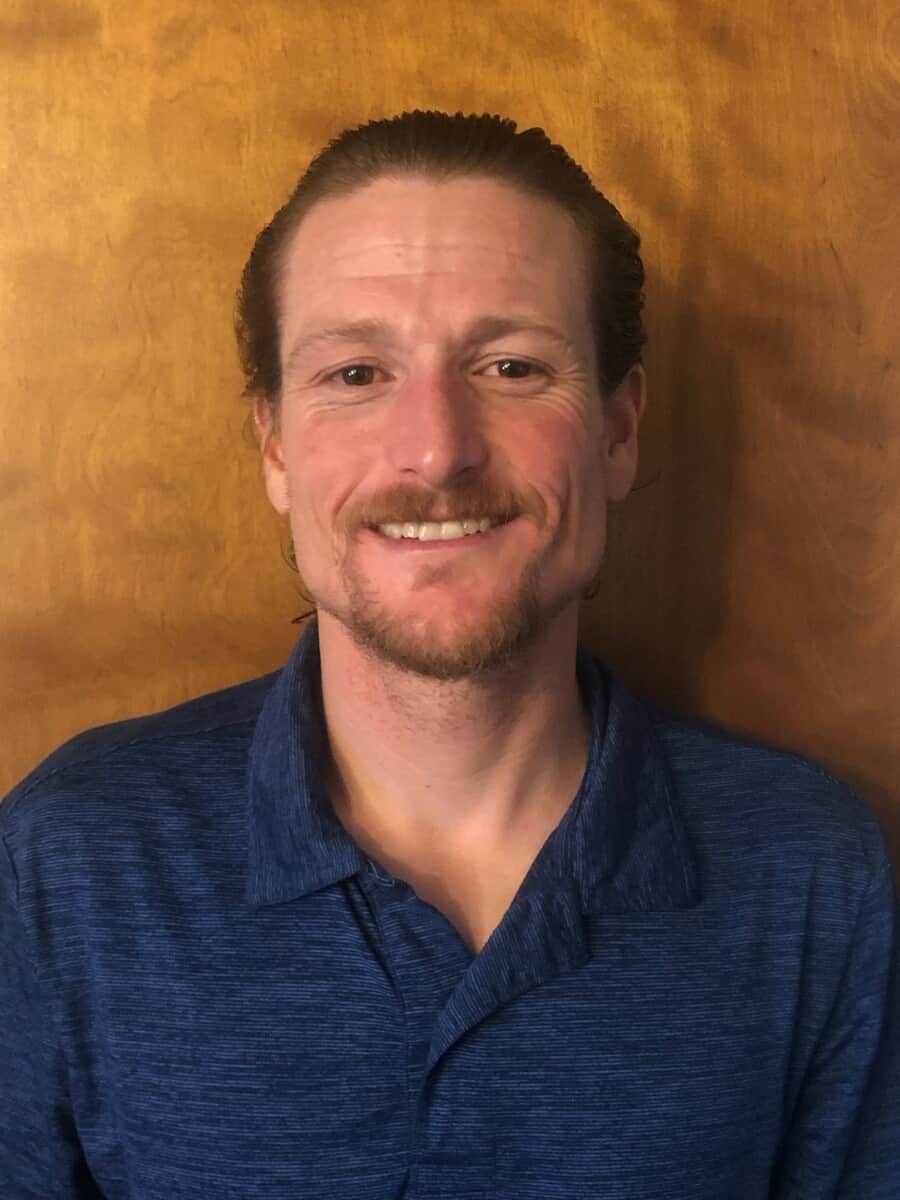 A man with short, slicked-back hair and a goatee is smiling at the camera. He is wearing a blue polo shirt and standing against a rustic wooden background, exuding confidence.
