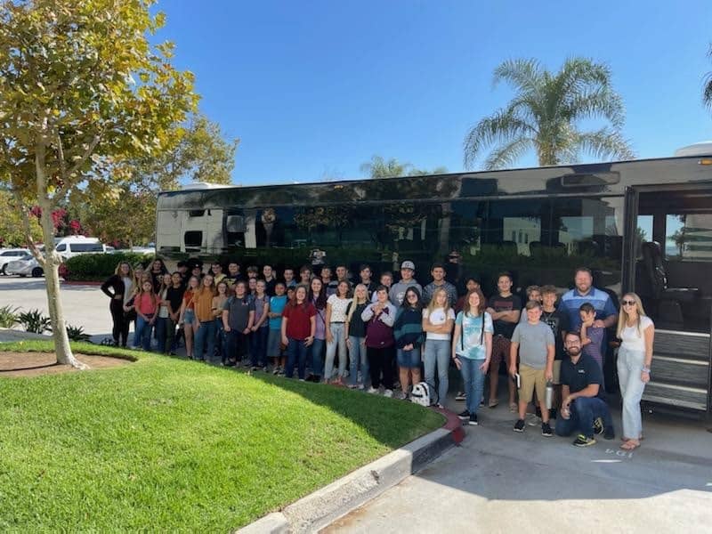 A group of engaging teenagers posing in front of a large bus on a sunny day with palm trees and a clear blue sky in the background.