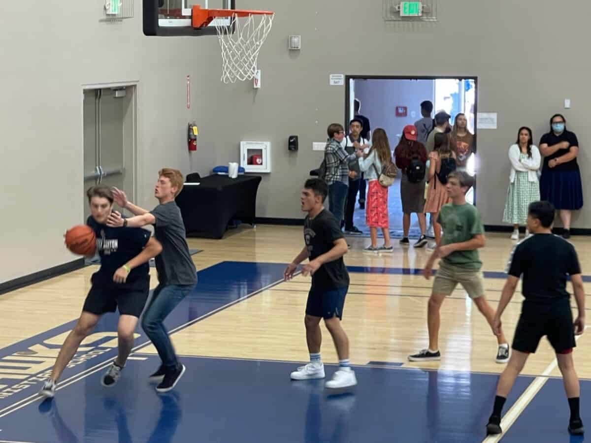 Teenagers playing basketball indoors, engaging in a fun game while others watch and chat in the background.
