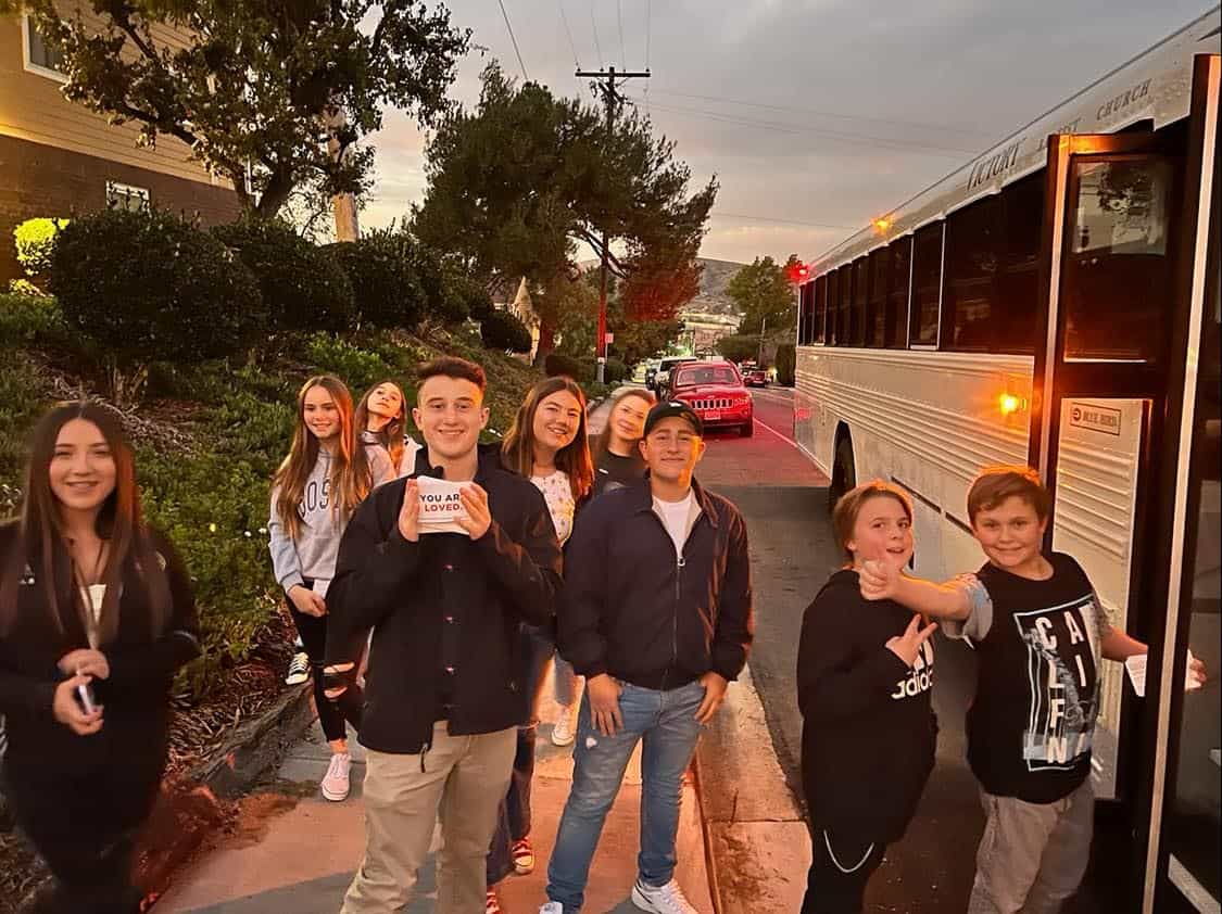 A youth group stands near a bus at dusk. Some are smiling, and one person holds a small sign that reads, "You are loved.