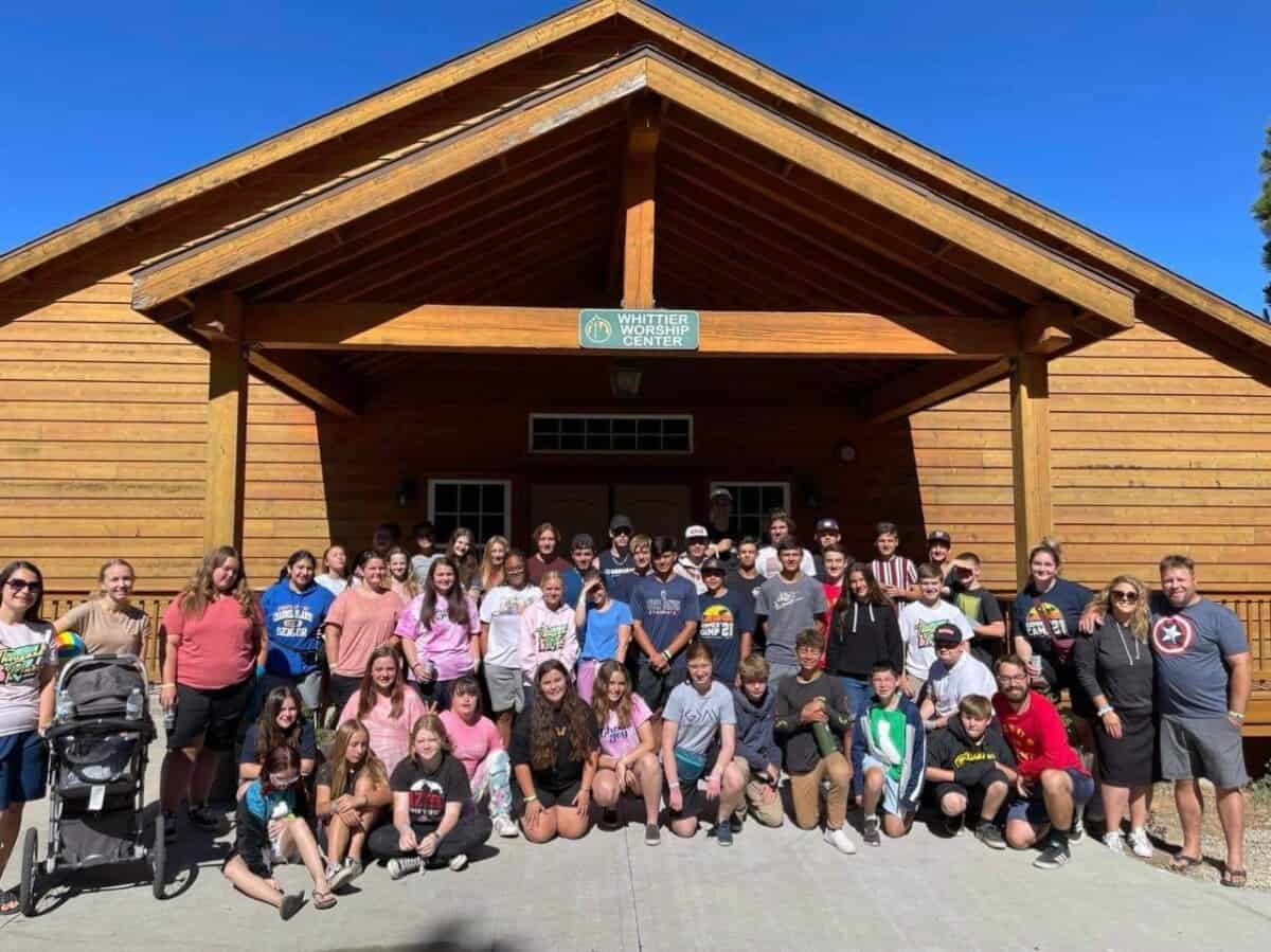 A large group of people, including children and adults, stand together in front of a wooden building under a clear blue sky. A sign reading "Writers Center" is visible above the entrance, creating an engaging atmosphere.