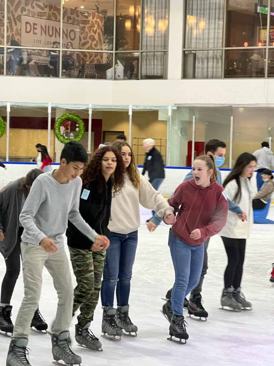 A group of teenagers ice skating indoors, holding hands for support. Some appear to be concentrating on maintaining balance on the ice. There is a restaurant in the background.