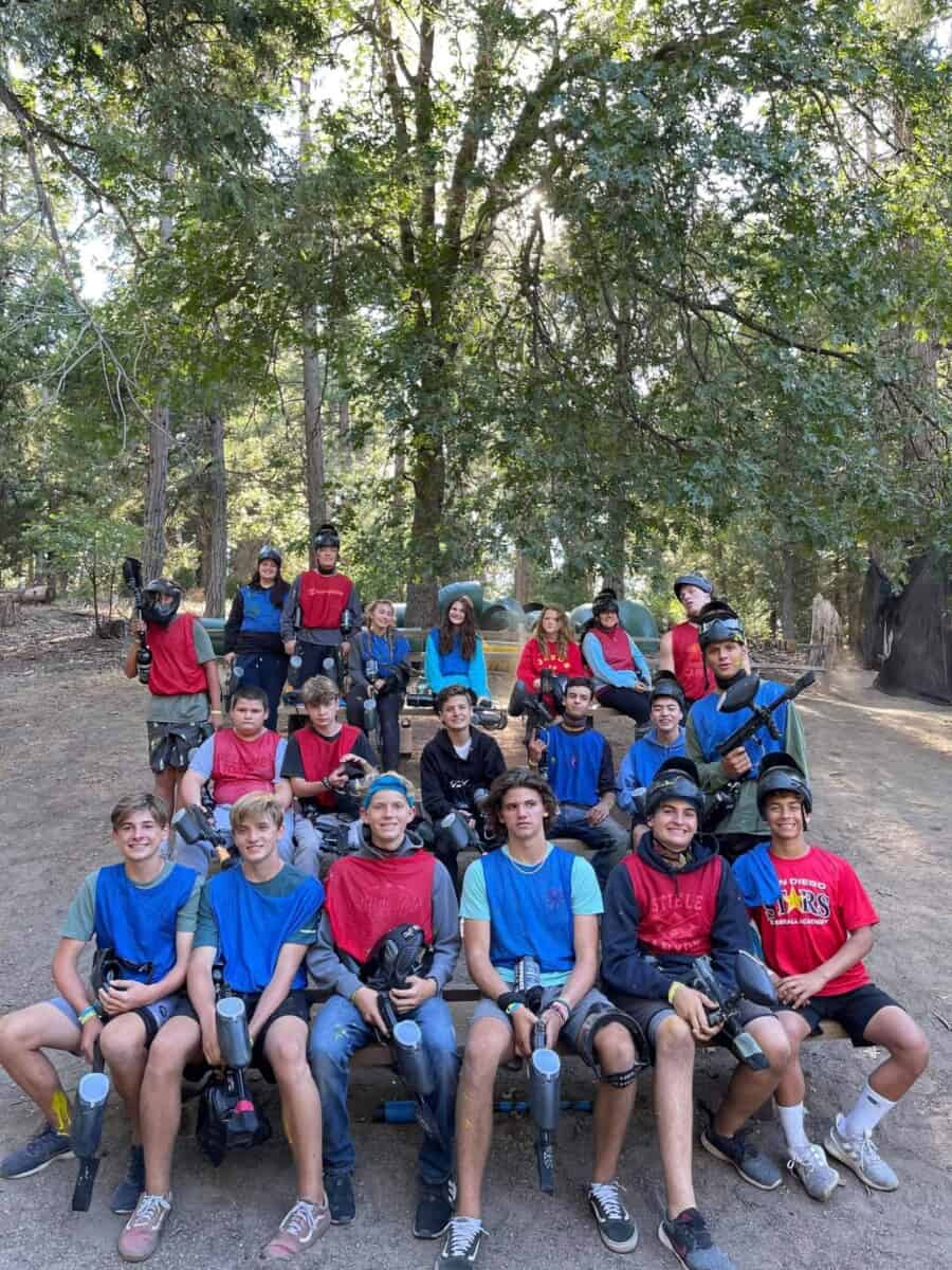 A youth group wearing protective gear and holding paintball guns pose for a fun group photo outdoors, with trees in the background. Some are seated while others stand behind them.