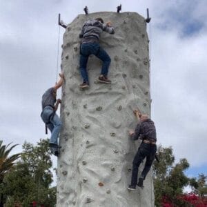 Three climbers scale a vertical rock wall outdoors under a cloudy sky, their determination evident in each move.