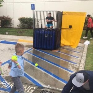 A young boy confidently throws a ball at a dunk tank target, aiming to dunk the person inside. The lively scene unfolds in a parking lot with accessible spaces, surrounded by greenery and a building in the background.