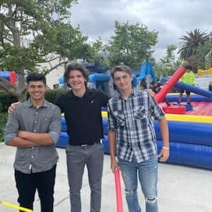 Three young men stand smiling in front of an inflatable obstacle course on a cloudy day, with trees and a bustling crowd visible in the background.