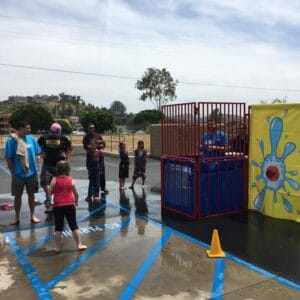 A child throws a ball towards a dunk tank while a man sits above the water inside, and several spectators watch nearby in an outdoor setting, eagerly anticipating the splash.